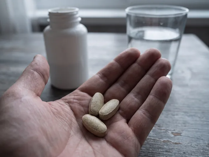 Open palm holding large vitamin tablets in front of a plain supplement bottle and a glass of water